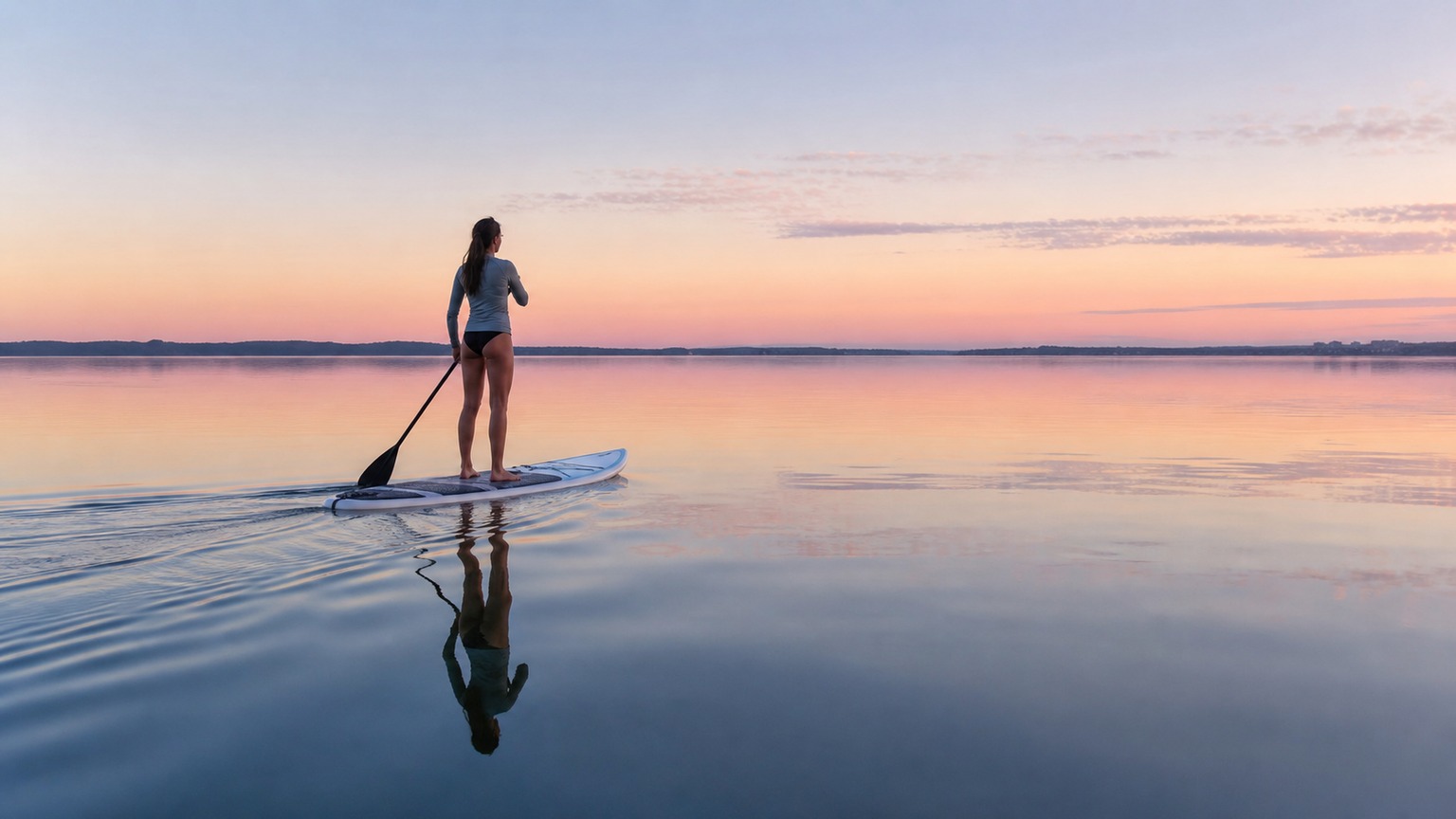 Woman stand-up paddleboarding on perfectly flat mirror-calm Choctawhatchee Bay at sunrise in Destin Florida, pink sky reflected in the still water